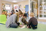 Eine Frau liest mit zwei Kindern in einem Spielraum. Alle schauen interessiert in ein Bilderbuch.
