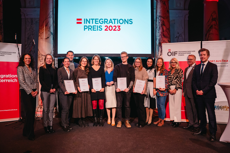 Gruppenfoto von Preisträgern des Integrationspreises 2023 mit Zertifikaten, vor einer Bildschirmpräsentation.