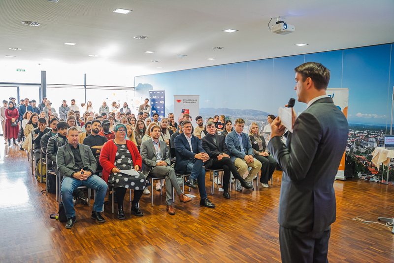Ein Redner spricht vor einem Publikum in einem modernen Raum. Viele Zuhörer hören aufmerksam zu.