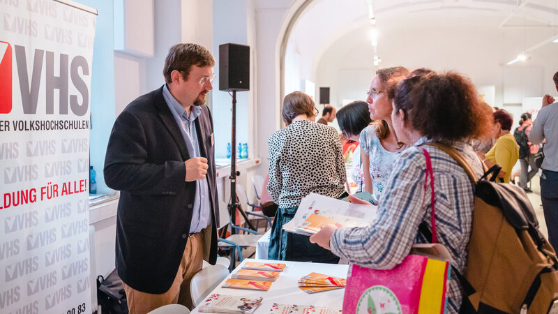 Eine Person steht am Informationsstand der VHS und spricht mit Besuchern über Bildungsangebote.