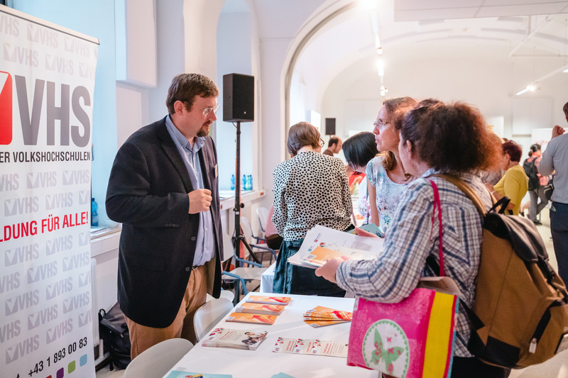 Eine Person steht am Informationsstand der VHS und spricht mit Besuchern über Bildungsangebote.