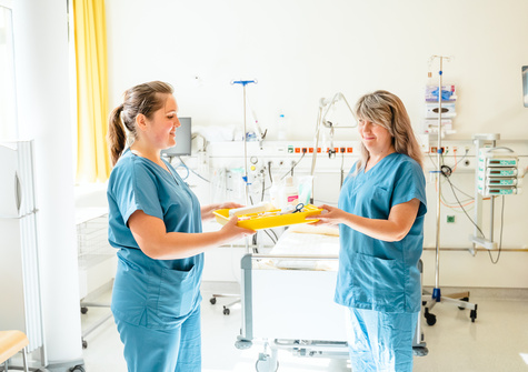 Zwei Krankenschwestern in blauen Einheiten reichen sich ein Tablett in einem Krankenhauszimmer.
