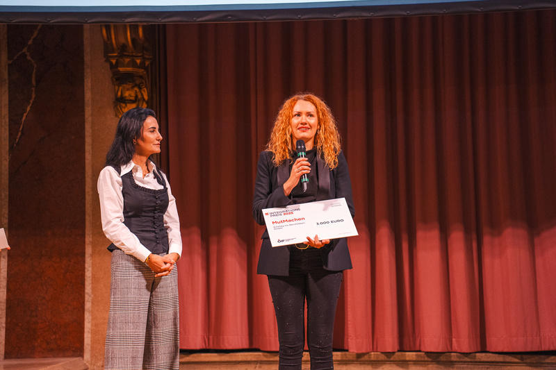 Zwei Frauen auf einer Bühne, eine hält ein Mikrophon und einen Scheck in der Hand.