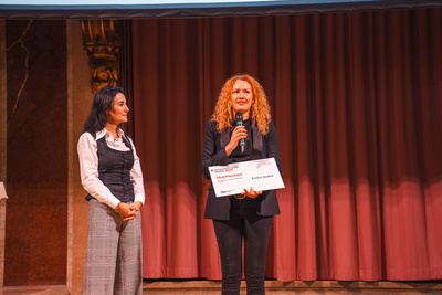 Zwei Frauen auf einer Bühne, eine hält ein Mikrophon und einen Scheck in der Hand.