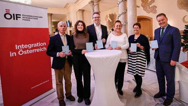 Sechs Personen halten Buchstaben und stehen an einem Stehtisch vor einem Plakat zur Integration in Österreich.