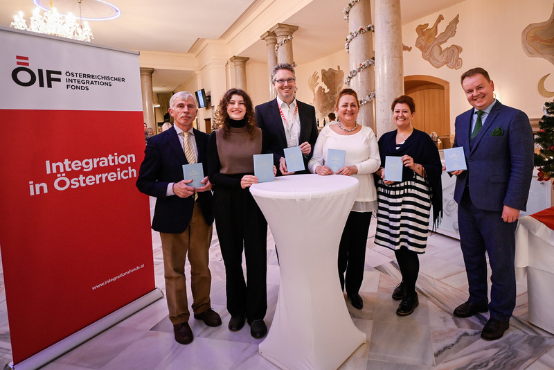 Sechs Personen halten Buchstaben und stehen an einem Stehtisch vor einem Plakat zur Integration in Österreich.