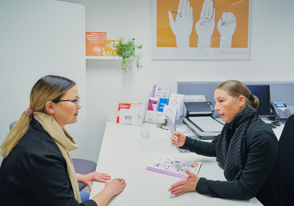 Zwei Frauen unterhalten sich an einem Schreibtisch in einem Büro. Eine Frau hält eine Broschüre.