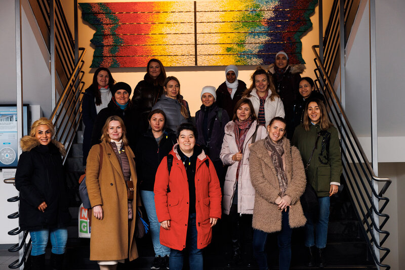 Eine Gruppe von 19 Frauen posiert lächelnd auf einer Treppe vor einem bunten Wandbild.