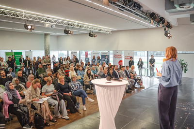 Eine Rednerin präsentiert vor einer großen Menschenmenge in einem Veranstaltungsraum.