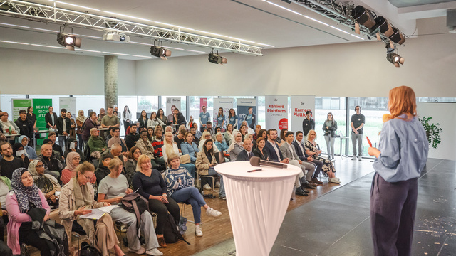 Eine Rednerin präsentiert vor einer großen Menschenmenge in einem Veranstaltungsraum.
