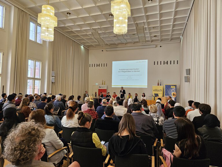 Ein Innenraum mit einer Vielzahl von Menschen, die an einer Veranstaltung teilnehmen. Auf der Bühne stehen mehrere Rednerinnen und Redner, im Hintergrund ist eine Präsentationsleinwand sichtbar. Die Atmosphäre ist informativ und ansprechend.