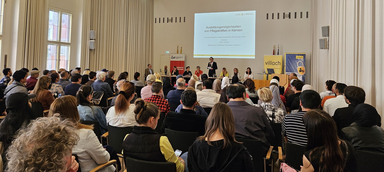 Ein Innenraum mit einer Vielzahl von Menschen, die an einer Veranstaltung teilnehmen. Auf der Bühne stehen mehrere Rednerinnen und Redner, im Hintergrund ist eine Präsentationsleinwand sichtbar. Die Atmosphäre ist informativ und ansprechend.