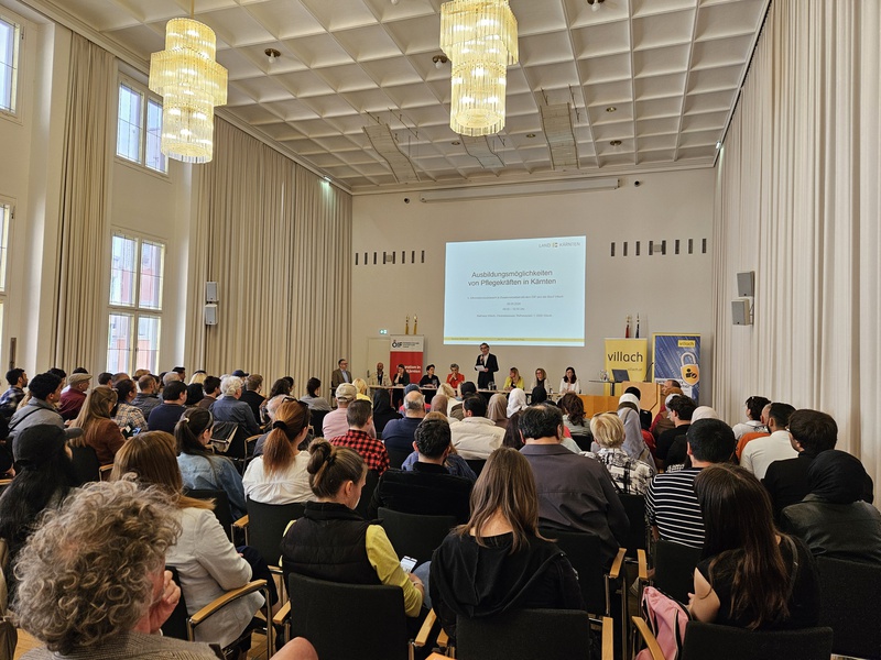 Ein Innenraum mit einer Vielzahl von Menschen, die an einer Veranstaltung teilnehmen. Auf der Bühne stehen mehrere Rednerinnen und Redner, im Hintergrund ist eine Präsentationsleinwand sichtbar. Die Atmosphäre ist informativ und ansprechend.