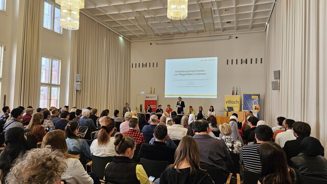 Ein Innenraum mit einer Vielzahl von Menschen, die an einer Veranstaltung teilnehmen. Auf der Bühne stehen mehrere Rednerinnen und Redner, im Hintergrund ist eine Präsentationsleinwand sichtbar. Die Atmosphäre ist informativ und ansprechend.