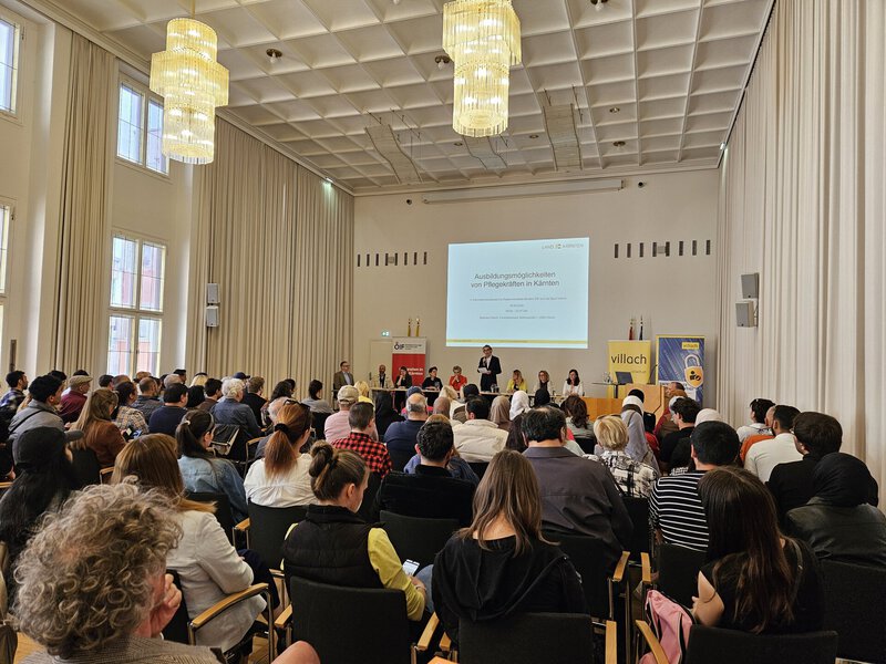 Ein Innenraum mit einer Vielzahl von Menschen, die an einer Veranstaltung teilnehmen. Auf der Bühne stehen mehrere Rednerinnen und Redner, im Hintergrund ist eine Präsentationsleinwand sichtbar. Die Atmosphäre ist informativ und ansprechend.