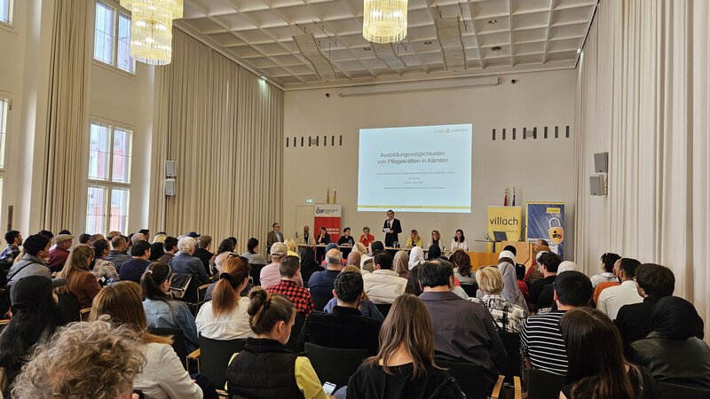 Ein Innenraum mit einer Vielzahl von Menschen, die an einer Veranstaltung teilnehmen. Auf der Bühne stehen mehrere Rednerinnen und Redner, im Hintergrund ist eine Präsentationsleinwand sichtbar. Die Atmosphäre ist informativ und ansprechend.