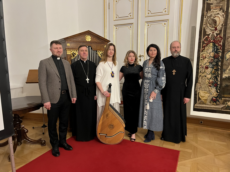 Sechs Personen in formeller Kleidung stehen nebeneinander, eine hält eine Bandura. Roter Teppich im Hintergrund.