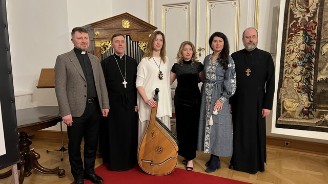 Sechs Personen in formeller Kleidung stehen nebeneinander, eine hält eine Bandura. Roter Teppich im Hintergrund.