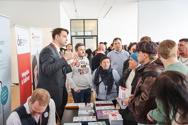 Eine Gruppe von Menschen steht um einen Tisch, an dem Informationsmaterialien ausgelegt sind. Ein Mann spricht mit den Anwesenden, während andere aufmerksam zuhören. Im Hintergrund sind Banner von Organisationen zu sehen.