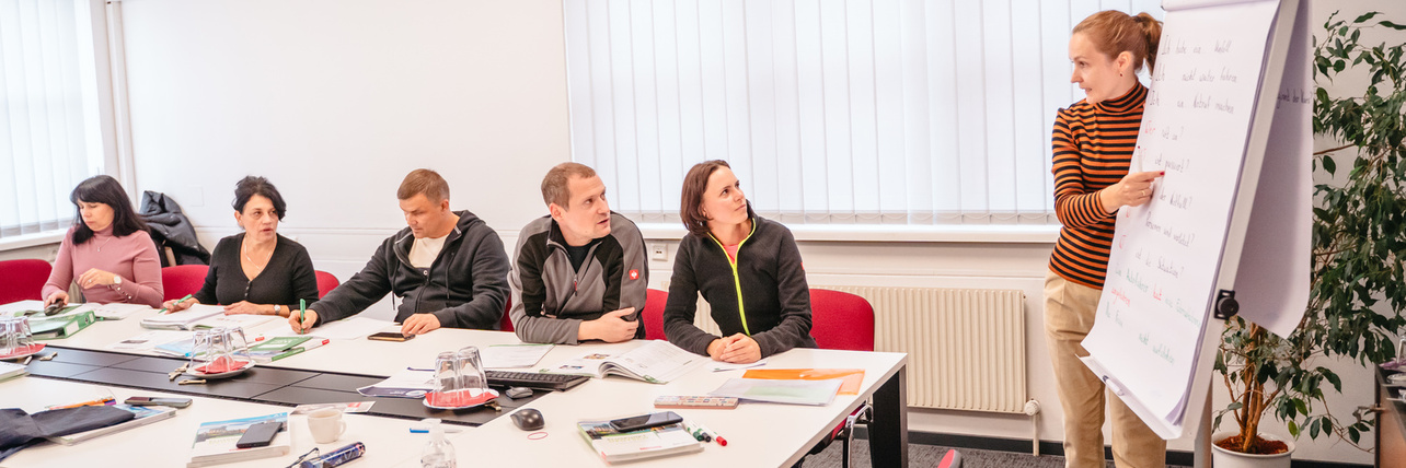 Eine Person präsentiert an einem Flipchart während einer Besprechung mit Teilnehmern am Tisch.