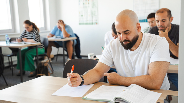 Ein Mann mit Glatze sitzt an einem Tisch und studiert ein Buch, während er mit einem Stift Notizen macht. Im Hintergrund sind weitere Personen zu sehen, die ebenfalls lernen. Der Raum ist hell und modern eingerichtet.