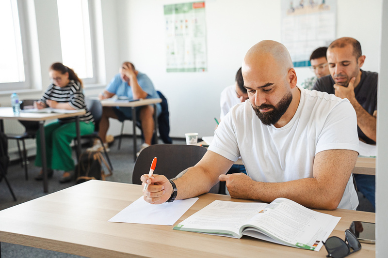 Ein Mann mit Glatze sitzt an einem Tisch und studiert ein Buch, während er mit einem Stift Notizen macht. Im Hintergrund sind weitere Personen zu sehen, die ebenfalls lernen. Der Raum ist hell und modern eingerichtet.