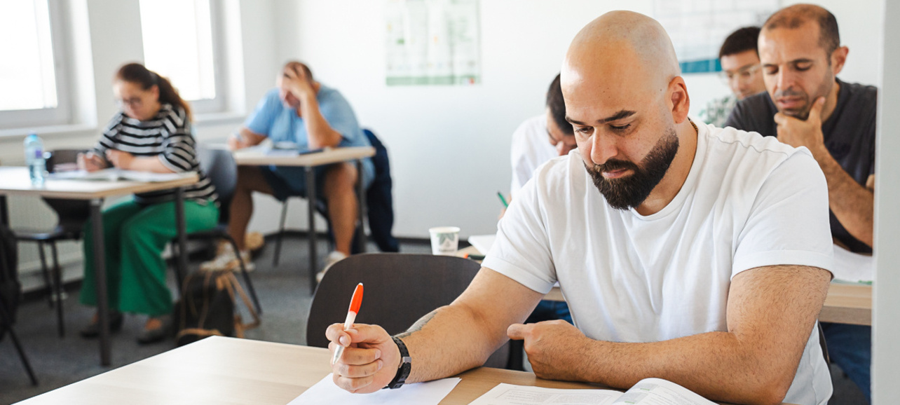 Ein Mann mit Glatze sitzt an einem Tisch und studiert ein Buch, während er mit einem Stift Notizen macht. Im Hintergrund sind weitere Personen zu sehen, die ebenfalls lernen. Der Raum ist hell und modern eingerichtet.