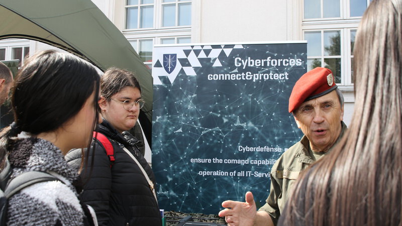 Ein militärischer Offizier in Uniform mit rotem Barett spricht mit einer Gruppe von Menschen vor einem Banner, das Informationen über Cyberabwehr enthält. Im Hintergrund ist ein Zelt sichtbar, während die Personen aufmerksam zuhören.