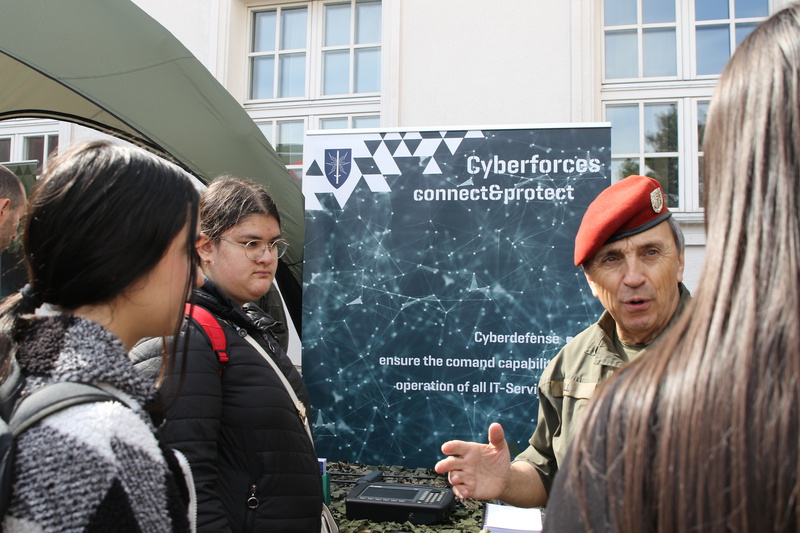 Ein militärischer Offizier in Uniform mit rotem Barett spricht mit einer Gruppe von Menschen vor einem Banner, das Informationen über Cyberabwehr enthält. Im Hintergrund ist ein Zelt sichtbar, während die Personen aufmerksam zuhören.