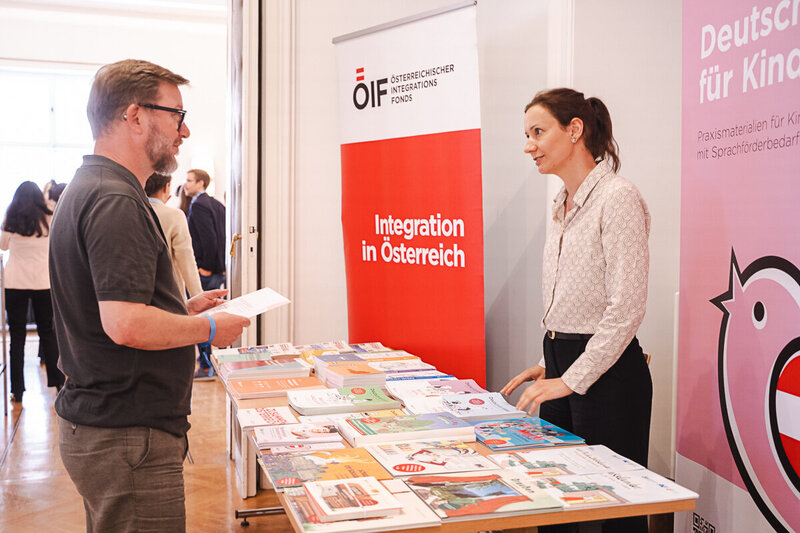 Eine Frau steht an einem Tisch mit verschiedenen Informationsmaterialien, während ein Mann ihr Fragen stellt. Im Hintergrund sind weitere Personen zu sehen. Ein Banner mit der Aufschrift "Integration in Österreich" ist deutlich erkennbar.