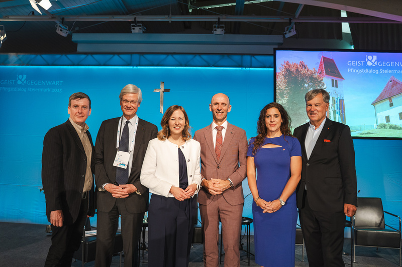 Sechs Personen stehen gemeinsam auf einer Bühne, vor einem blauen Hintergrund mit einem religiösen Symbol.