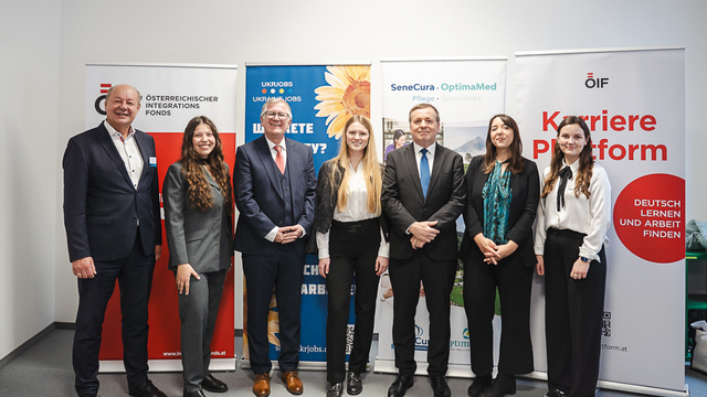 Eine Gruppe von sieben Personen steht in einem Raum, umgeben von Bannern verschiedener Organisationen.