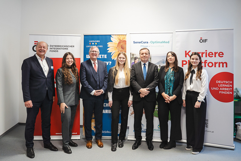 Eine Gruppe von sieben Personen steht in einem Raum, umgeben von Bannern verschiedener Organisationen.