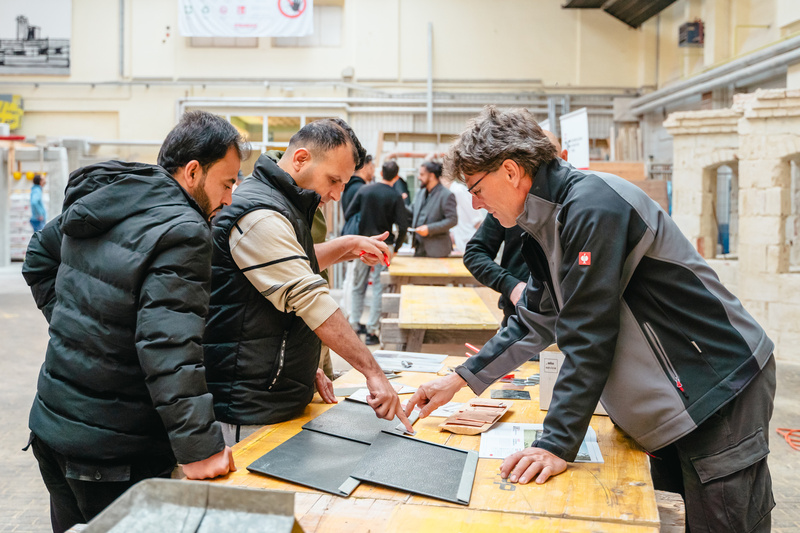 Vier Personen diskutieren an einem Tisch über Materialien in einer Werkstattumgebung.