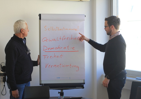 Zwei Personen stehen vor einem Flipchart mit Werten wie Selbstbestimmung, Demokratie und Freiheit.