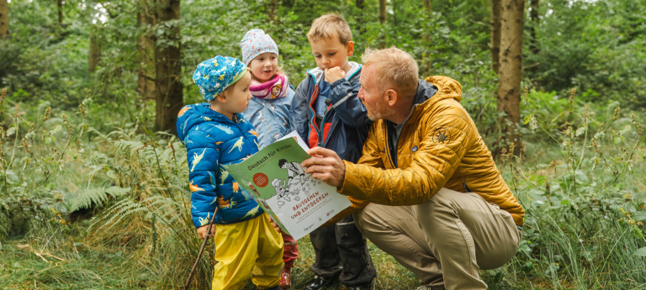 Ein Mann zeigt Kindern in einem Wald ein Buch und erklärt ihnen etwas darüber.