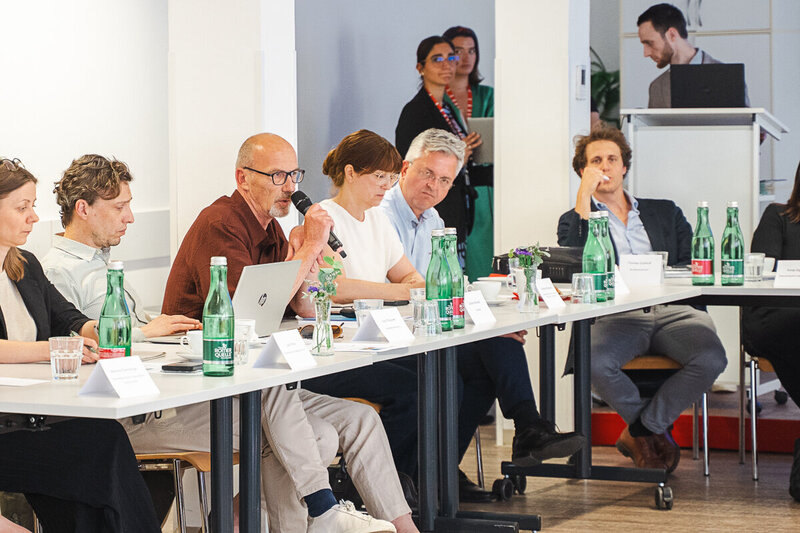 Eine Gruppe von Fachleuten sitzt um einen Tisch in einem Besprechungsraum. Einige Teilnehmer sprechen, während andere zuhören. Auf dem Tisch stehen Wasserflaschen und Notizen. Im Hintergrund ist ein Präsentationsbereich sichtbar.
