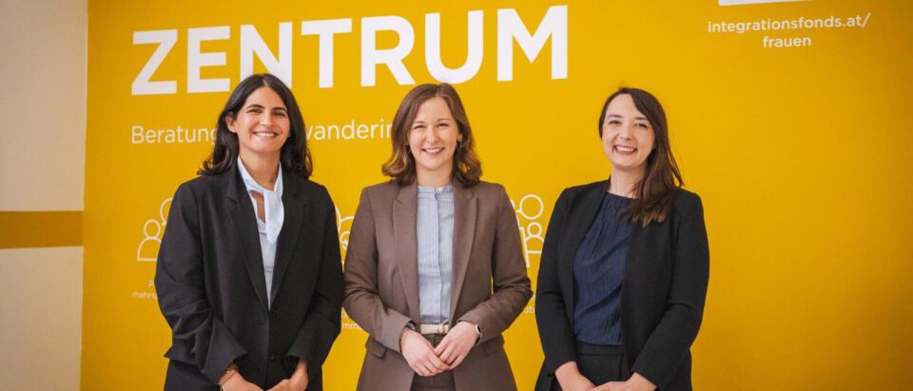 Drei Frauen stehen vor einer gelben Wand mit dem Schriftzug "ZENTRUM". Sie lächeln und sind in formeller Kleidung gekleidet. Die Wand enthält Informationen über Beratung für Migrantinnen.
