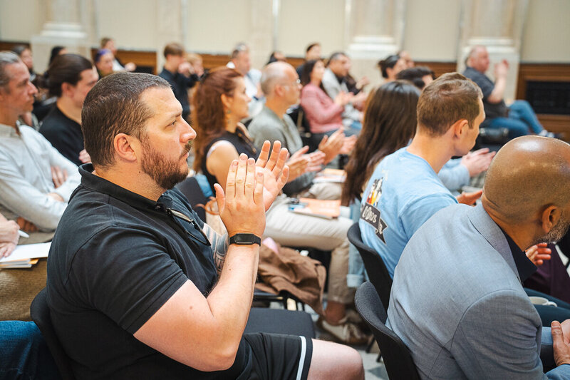 Eine Gruppe von Menschen sitzt in einem Raum und applaudiert. Der Fokus liegt auf einem Mann im Vordergrund, der in einem schwarzen T-Shirt sitzt und applaudiert. Die Zuschauer wirken aufmerksam und engagiert.