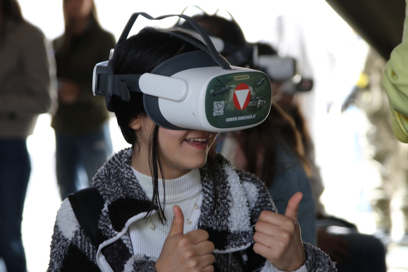 Ein Mädchen trägt eine VR-Brille und lächelt, während sie beiden Daumen nach oben zeigt. Im Hintergrund sind weitere Personen zu sehen, die ebenfalls an einer Veranstaltung teilnehmen.