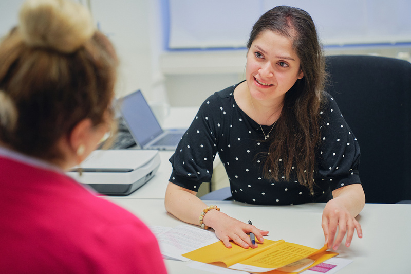 Zwei Frauen führen ein Gespräch am Tisch, eine schreibt Notizen auf einem gelben Blatt.