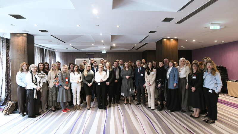 Gruppenfoto von etwa 40 Personen, die in einem modernen Raum zusammenstehen. Die Teilnehmerinnen und Teilnehmer tragen unterschiedliche Kleidung, einige haben Namensschilder. Der Raum ist hell erleuchtet mit großen Fenstern und einem eleganten Bodenbelag.