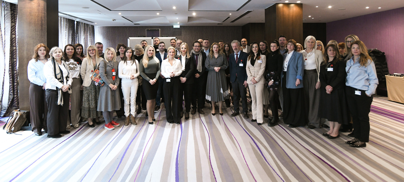 Gruppenfoto von etwa 40 Personen, die in einem modernen Raum zusammenstehen. Die Teilnehmerinnen und Teilnehmer tragen unterschiedliche Kleidung, einige haben Namensschilder. Der Raum ist hell erleuchtet mit großen Fenstern und einem eleganten Bodenbelag.