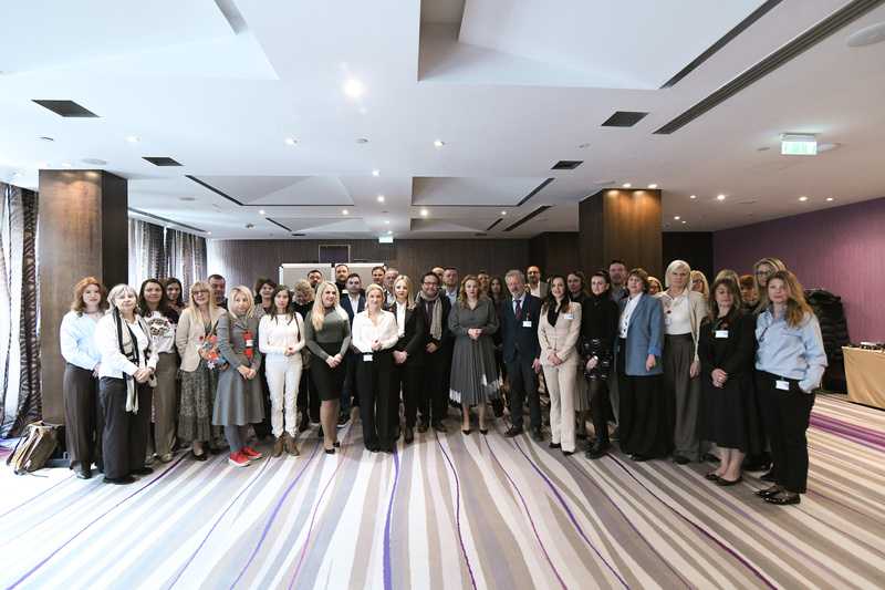 Gruppenfoto von etwa 40 Personen, die in einem modernen Raum zusammenstehen. Die Teilnehmerinnen und Teilnehmer tragen unterschiedliche Kleidung, einige haben Namensschilder. Der Raum ist hell erleuchtet mit großen Fenstern und einem eleganten Bodenbelag.