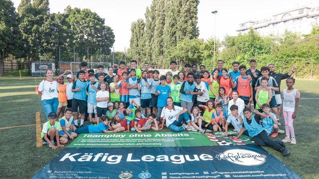 Eine Gruppe von Kindern und Jugendlichen posiert lächelnd auf einem Fußballfeld vor einem Banner.