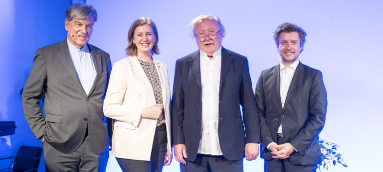 Vier Personen stehen zusammen auf einer Bühne, lächelnd, vor einem blauen Hintergrund.