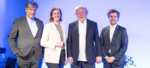 Vier Personen stehen zusammen auf einer Bühne, lächelnd, vor einem blauen Hintergrund.