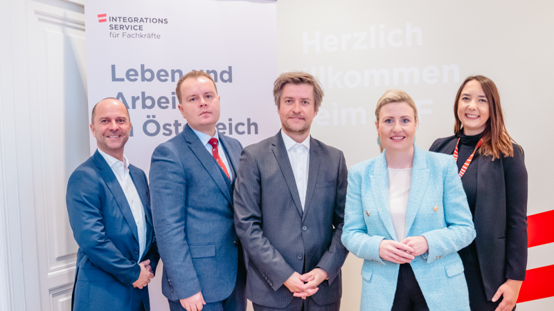 Fünf Personen stehen lächelnd vor einem Willkommensbanner für den Integrationsservice in Österreich.