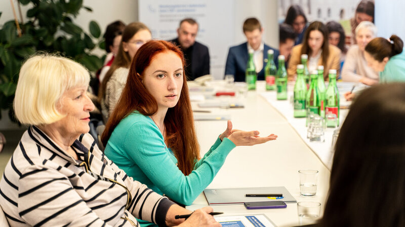 Zwei Frauen diskutieren an einem Tisch, umgeben von Teilnehmern und Getränken.
