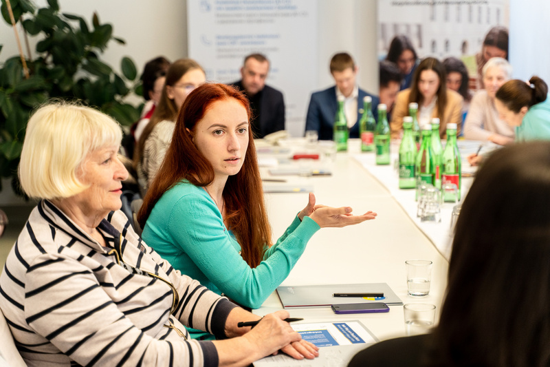 Zwei Frauen diskutieren an einem Tisch, umgeben von Teilnehmern und Getränken.